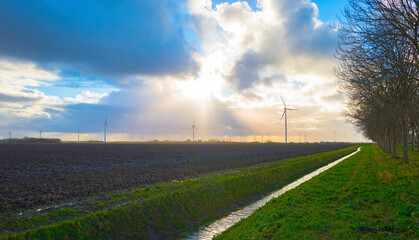 Wind turbines on the horizon of an agricultural field along a ditch in bright sunlight in winter,...