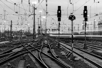 Railway infrastructure at Frankfurt main station, Germany. Black and white panorama. Technology with signals, crossings, catenary, tracks, switches at twilight. Blurred fast train lights in motion. 