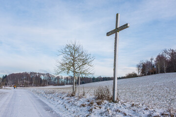 Teilweise verschneites Feldkreuz mit Flechtenbesatz an Feldweg auf der schwäbischen Alb vor blauem Himmel mit Schleierwolken