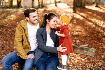 Little girl touches dad sitting with mom on bench in autumn forest
