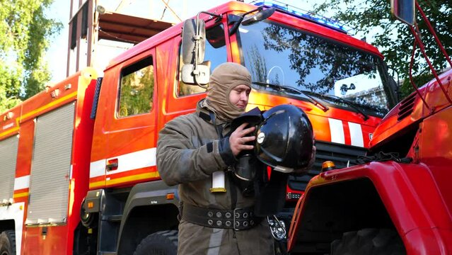 Male firefighter in uniform taking off helmet stands near a fire engine. Young fireguard removing equipment from himself after fire against background of a big red truck. Concept of saving lives
