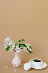 Beautiful branches of a blooming apple tree in a pink vase with a cup of tea and marshmallows on a beige background with a copy space