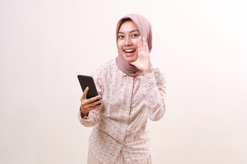 Fototapeta premium Excited young asian muslim girl standing announcing something while holding a cell phone. Isolated on white