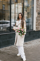 Stylish young woman with a bouquet of tulips outdoors and holding a paper cup with coffee in her hands. Portrait of a spring street lifestyle