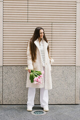 Fototapeta premium Young woman in stylish light coloured clothes strolls through the streets of the city with a bouquet of tulips