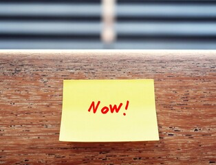 A yellow note pad with red written message text NOW ,stick on the wood hand rail  with blurred...