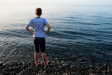 Teen boy standing in sea on pebble beach looking at transparent clear water. Teenger on vacation...