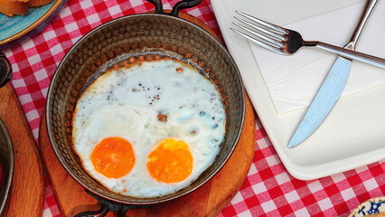 wo perfectly fried eggs sizzle in a skillet, set on a table with a charming red checkered tablecloth. A plate with a fork and knife awaits, ready to indulge in this delightful breakfast delight