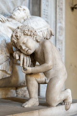Fototapeta na wymiar Close-up on marble statue of little naked boy kneeling in sorrow while holding the hand of a dead old man laying on a bed