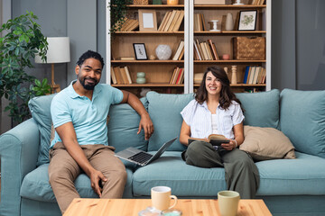 Cheerful millennial mixed couple spending time with laptop and book at home, happy young interracial lovers sitting on sofa with computer, freelance working or shopping online together and reading
