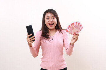 Wow excited young asian girl holding cell phone and Indonesia banknotes while looking at the camera. Isolated on white