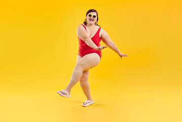 Portrait of overjoyed excited happy funny fat woman in red swimsuit and sunglasses having fun and dancing isolated on a studio yellow background. Summer holiday trip and vacation concept. - obrazy, fototapety, plakaty