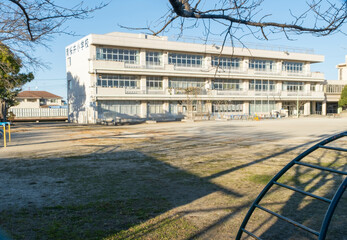 The exterior of a Japanese elementary school building and the empty dirt playing field in front of it on a cold January afternoon. 