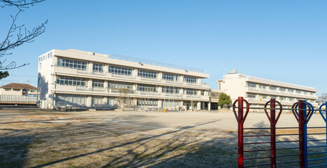 The exterior of a Japanese elementary school building and the empty dirt playing field in front of...