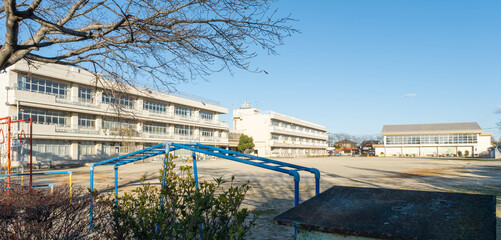The exterior of a Japanese elementary school building and the empty dirt playing field in front of...