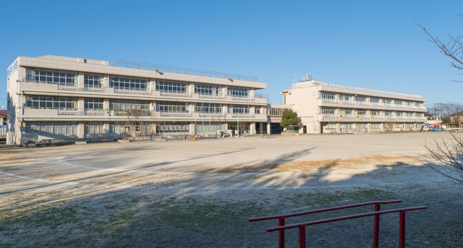 The exterior of a Japanese elementary school building and the empty dirt playing field in front of it on a cold January afternoon. 