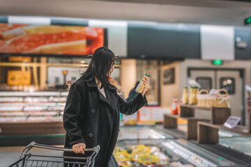 A young Asian woman is pushing a shopping cart in the food section of a supermarket, reading the...