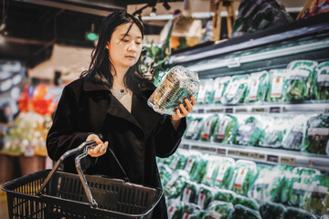 A young Asian woman with a shopping basket, standing next to a supermarket shelf, filming the...