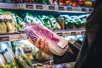 A young Asian woman with a shopping basket, standing next to a supermarket shelf, filming the purchase of fresh organic vegetables