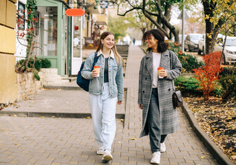 Naklejka premium Two multiracial female best friends walking city street with take away coffee, enjoying autumn walk