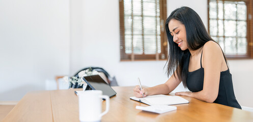 Young asian woman in casual wear writing note while sitting in living room at home.