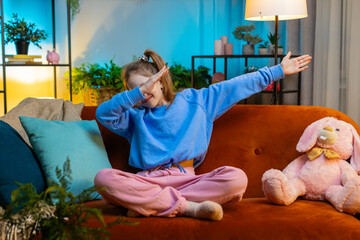 Trendy cheerful positive redhead girl having fun dancing, moving to rhythm, dabbing raising hands, making dub dance gesture. Teenager child kid at home in night evening play room sitting on couch