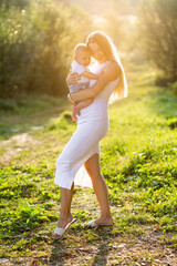 Happy young mother hug with her little baby son on sunshine warm spring or summer day. Beautiful sunset light in the apple garden or in the park. family concept