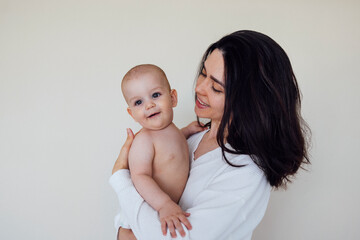 Young beautiful mother holds her cute baby in her arms. Pretty infant with his smiling brunette mom in white clothes on beige background.