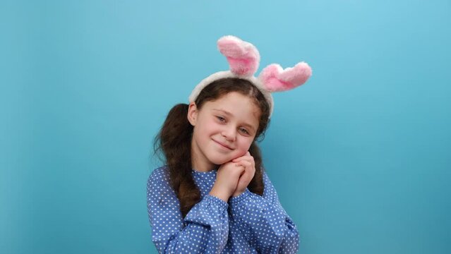 Portrait of funny smiling charming preteen girl child with pink fluffy bunny ears, happy looking at camera, posing isolated over plain blue color background wall in studio. Easter holiday concept