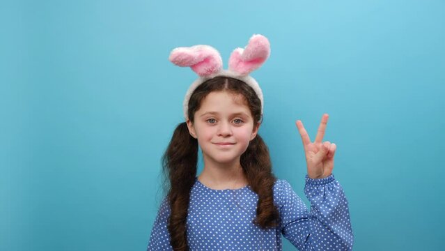 Portrait of adorable happy preteen girl kid wearing pink bunny ears, smiling cute child having fun, showing v sign to camera, posing isolated over blue color background wall in studio. Easter concept
