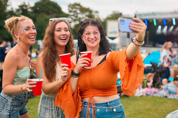 Three Female Friends Wearing Glitter Posing For Selfie At Summer Music Festival Holding Drinks