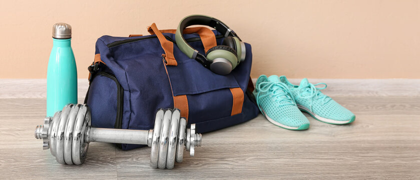 Sports bag with headphones, shoes , dumbbell and bottle near beige wall in gym