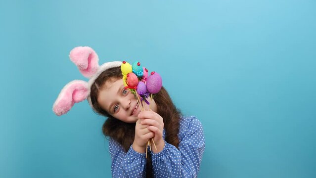 Portrait of funny happy little girl kid with Easter bunny ears and colorful small Easter eggs, playful looking at camera, posing isolated over blue color background wall in studio. Easter concept