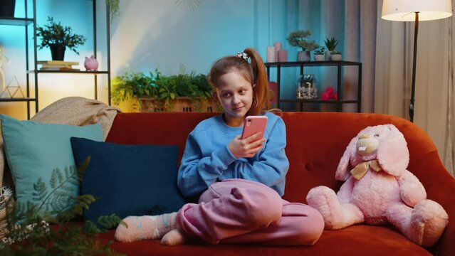 Young child girl texting share messages content on smartphone social media applications online, watching relax movie. Female teenager kid uses mobile phone at home in evening night room sits on sofa