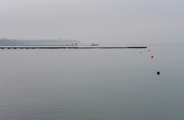 Grado sea during a winter day with a thick fog which makes the environment suggestive and mystical. Shot of the beach with people walking on the pier overlooking the sea in the distance. 