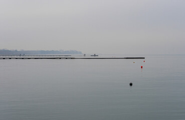 Grado sea during a winter day with a thick fog which makes the environment suggestive and mystical. Shot of the beach with people walking on the pier overlooking the sea in the distance. 