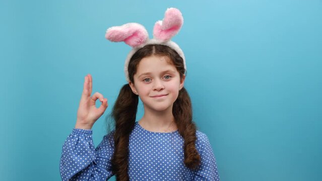 Portrait of beautiful smiling preteen girl kid wearing pink bunny ears and casual dress, happy looking at camera, showing okay sign with fingers, posing isolated over blue studio background wall
