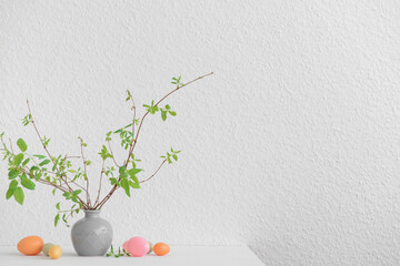 Vase with tree branches and painted Easter eggs on table near white wall