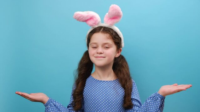 Portrait of cute preteen girl kid in pink bunny fluffy ears holds copy space with palms, has good mood, isolated over blue studio background wall with empty space for your text. Easter holiday concept