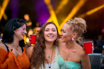 Three Female Friends Wearing Glitter Having Fun At Summer Music Festival Holding Drinks