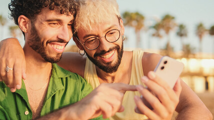 Close-up of young gay couple sitting embracing, smiling and laughing have fun with mobile phone...