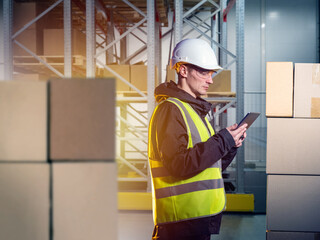 Man warehouse worker. Guy is standing in storage hangar. Warehouse worker with electronic tablet. Supervisor works in storage facility. Boxes and multi-tiered shelving near storekeeper man