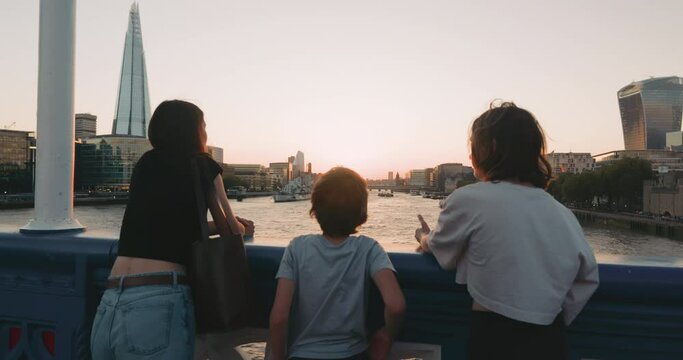 Young female and two teenage boy and girl admire beautiful sunset over the Thames river together.