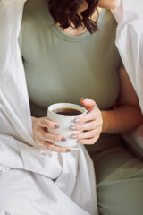 Relaxed positive young brunette woman in homewear sitting on top of unmade bed with cup of tea in her hands, slowly drinking aromatic hot beverage while enjoying spending free time alone at home. 