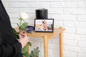 Woman holding rose flower, frame with picture of dog, collar and mortuary urn on table near white...