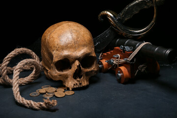 Human skull with sword, toy model of cannon, pirate hat and coins on black background