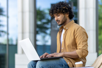 Concentrated and serious young Indian man using a laptop while sitting on a bench outside