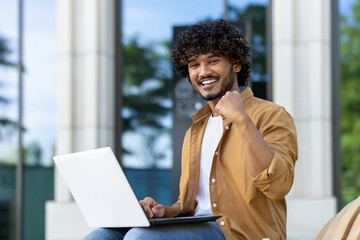 Portrait of a young Indian man who is happy for the camera showing a victory gesture with his hand, sitting on a bench on the street with a laptop