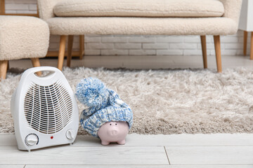 Electric fan heater, piggy bank and hat on floor. Heating season
