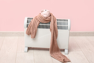 Electric convector heater, piggy bank and scarf near pink wall. Heating season